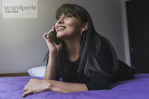 Lächelnde Frau  die auf dem Bett liegend mit einem Mobiltelefon spricht