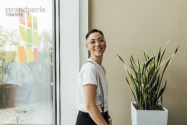 Lächelnde junge Geschäftsfrau mit Brille steht am Fenster