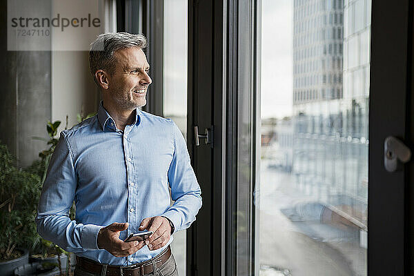 Lächelnder Geschäftsmann  der durch ein Fenster schaut  während er ein Mobiltelefon am Arbeitsplatz hält