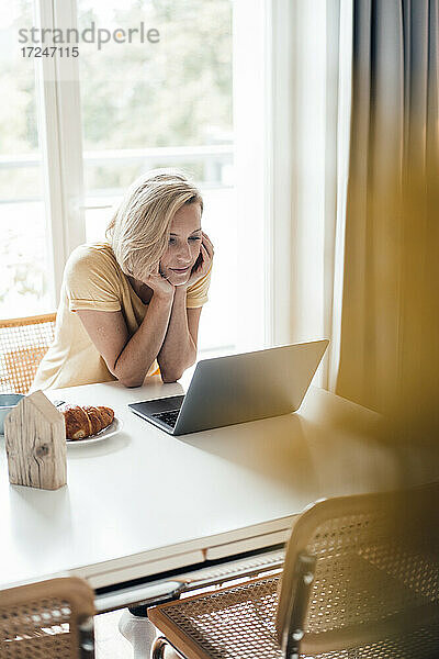 Berufstätige Frau mit Hand am Kinn  die zu Hause einen Laptop benutzt