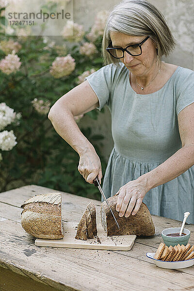 Reife Frau mit Brille schneidet Brot auf einem Tisch