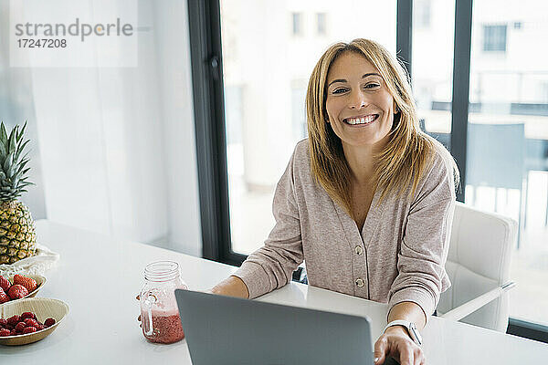 Lächelnde Frau im mittleren Erwachsenenalter mit Laptop auf dem Tisch zu Hause