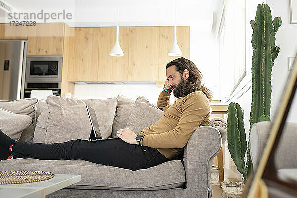 Mann mit langen Haaren  der einen Tablet-PC benutzt  während er sich auf dem Sofa zu Hause entspannt