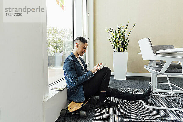 Unternehmer  der ein digitales Tablet benutzt  während er im Büro auf einem Skateboard sitzt