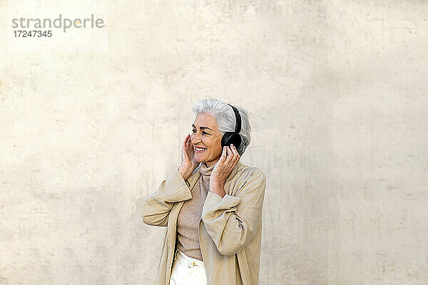Lächelnde reife Frau hört Musik über Kopfhörer vor einer Wand