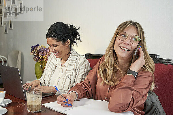 Lächelnde Geschäftsfrau  die wegschaut und mit einem Smartphone spricht  während sie mit einer Kollegin im Café sitzt