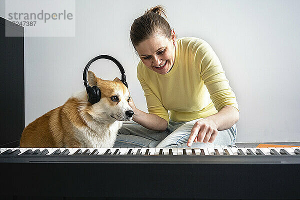 Lächelnde Frau mit Hund  die dem Hund Kopfhörer aufsetzt  während sie im Wohnzimmer Klavier übt