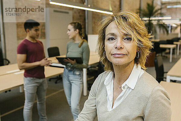 Reife Geschäftsfrau mit blondem Haar mit männlichen und weiblichen Mitarbeitern im Hintergrund im Büro