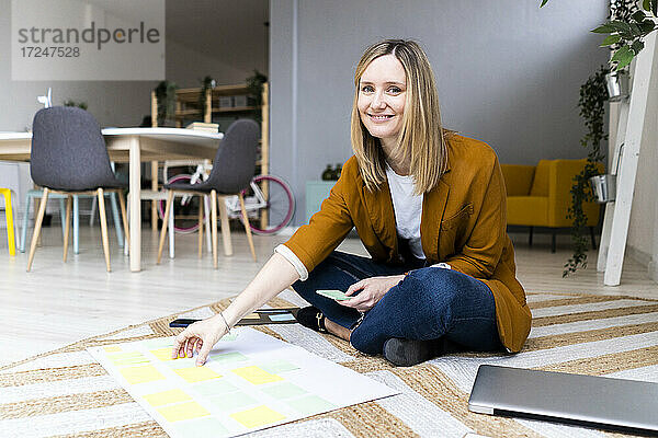 Lächelnde kreative Geschäftsfrau mit Haftnotizen auf Teppich im Büro sitzend