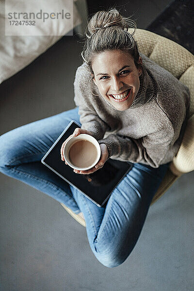 Lächelnde erwachsene Geschäftsfrau  die eine Getränkeschale hält  während sie mit einem digitalen Tablet in einem Café sitzt