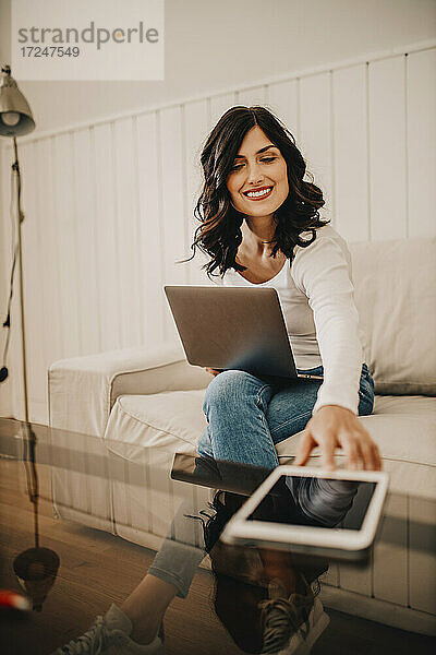 Freiberuflerin greift nach einem digitalen Tablet  während sie zu Hause am Laptop sitzt