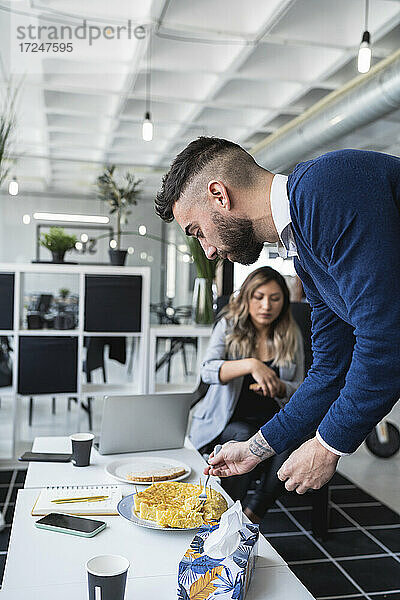 Geschäftsmann isst spanisches Omelett am Tisch mit einer Kollegin im Hintergrund im Büro