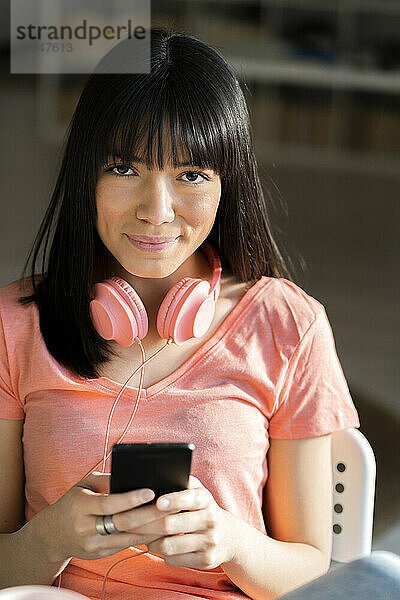 Lächelnde junge Frau mit Kopfhörern und Smartphone zu Hause