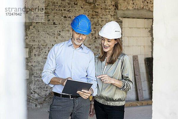 Weiblicher Bauherr und männlicher Architekt mit Schutzhelmen diskutieren auf einer Baustelle über ein digitales Tablet