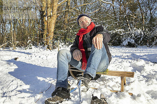 Glücklicher älterer Mann  der an einem sonnigen Tag auf einem Schlitten im Schnee sitzt
