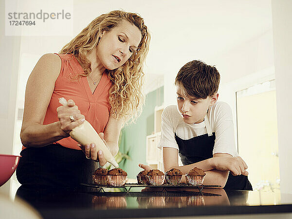 Sohn schaut Mutter an  die Muffins mit einem Spritzbeutel in der Küche glasiert