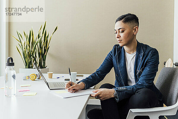 Unternehmer schreibt in Papier  während er mit Laptop im Büro sitzt