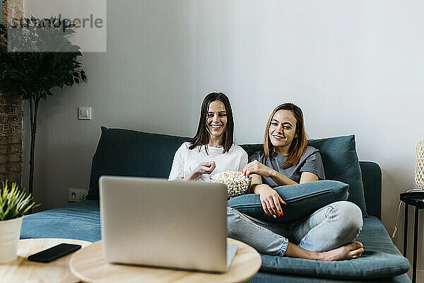 Lächelnde junge Freundinnen sehen sich einen Film auf dem Laptop an und essen Popcorn