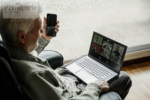 Älterer Mann mit Mobiltelefon  der einen Videoanruf über einen Laptop zu Hause führt