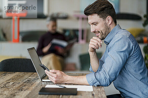 Männlicher Berufstätiger  der ein Smartphone neben einem Laptop im Büro benutzt