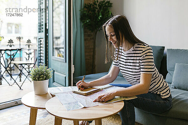 Lächelnde Frau schreibt eine Notiz in ihr Tagebuch  während sie am Tisch in einer Wohnung sitzt