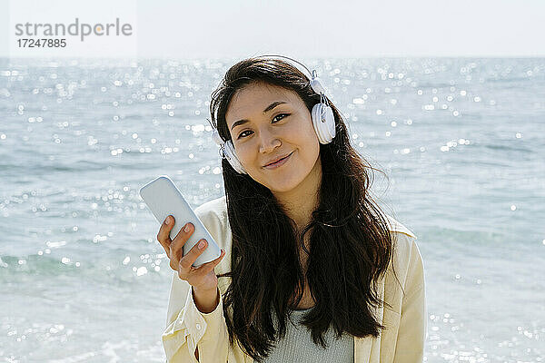 Lächelnde Frau mit Handy in der Hand  die an einem sonnigen Tag über Kopfhörer Musik hört