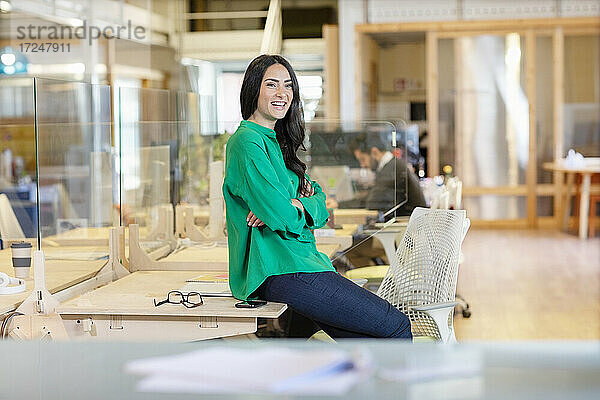 Lächelnde Unternehmerin  die wegschaut  während sie auf dem Schreibtisch im Büro sitzt