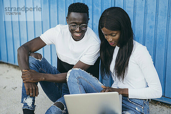 Frau benutzt Laptop  während sie mit ihrem Freund in der Nähe einer blauen Wand sitzt