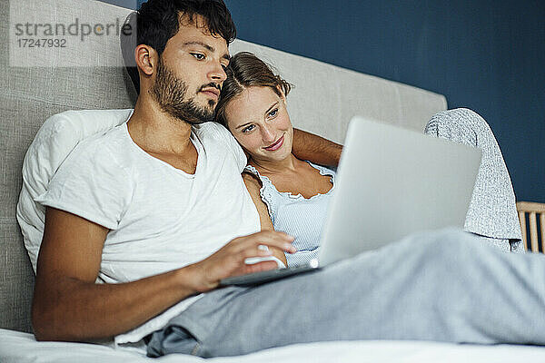 Junger Mann  der einen Laptop benutzt  während er neben einer Frau im Bett zu Hause sitzt
