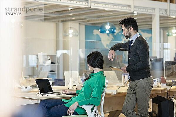 Junger Geschäftsmann mit digitalem Tablet  der hinter einer Kollegin mit Laptop im Büro steht
