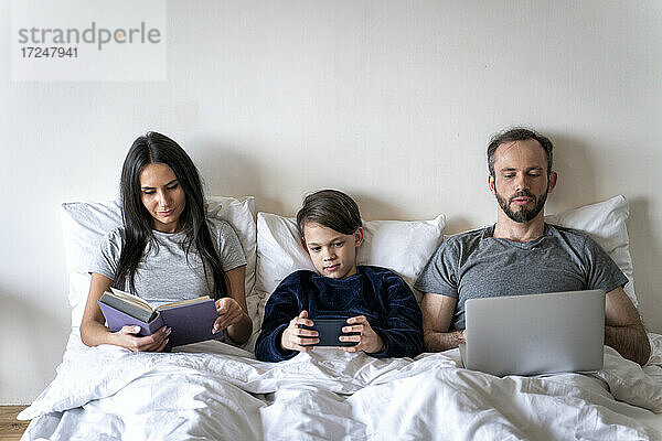 Frau liest ein Buch  während Vater und Sohn zu Hause im Bett drahtlose Technologien nutzen