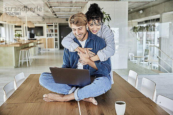 Lächelnde Frau umarmt Freund mit Laptop auf dem Tisch im Loft