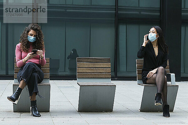 Frau mit Gesichtsmaske  die ein Mobiltelefon benutzt  während sie in sozialer Distanz mit einer Frau auf dem Gehweg sitzt