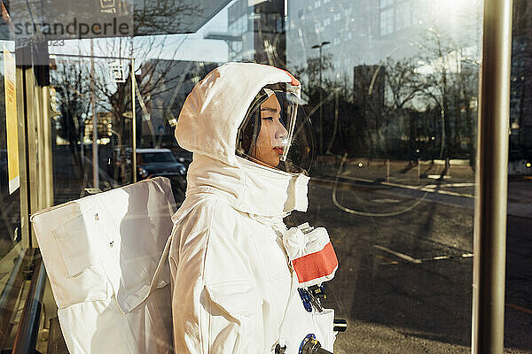 Weibliche Astronautin im Raumanzug an einer Bushaltestelle an einem sonnigen Tag durch Glas gesehen