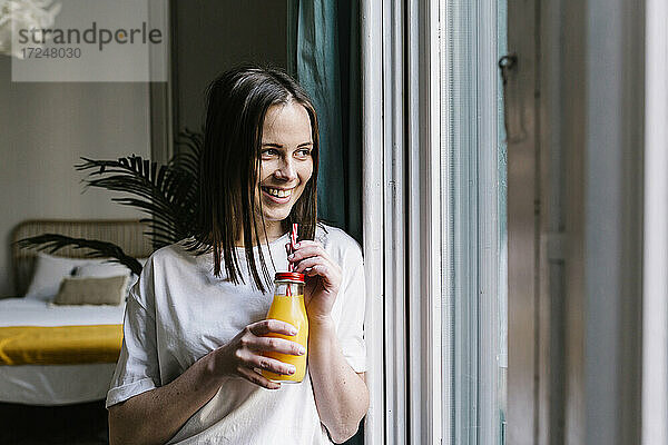 Lächelnde junge Frau schaut weg  während sie zu Hause am Fenster Orangensaft trinkt