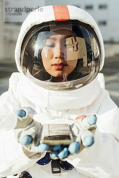 Weibliche Astronautin im Raumanzug schaut an einem sonnigen Tag auf das Smartphone in der Hand