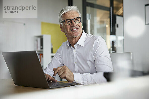 Lächelnder Geschäftsmann mit Laptop  der im Büro nachdenkt