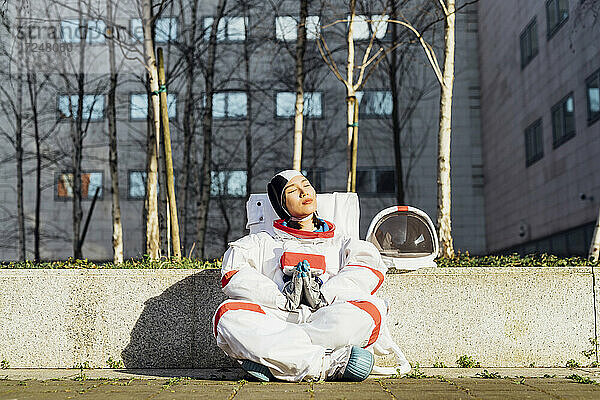 Weibliche Astronautin im Raumanzug  die mit verschränkten Händen an einer Stützmauer sitzt  an einem sonnigen Tag