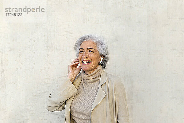 Glückliche Frau hört Musik über drahtlose Kopfhörer vor einer Wand