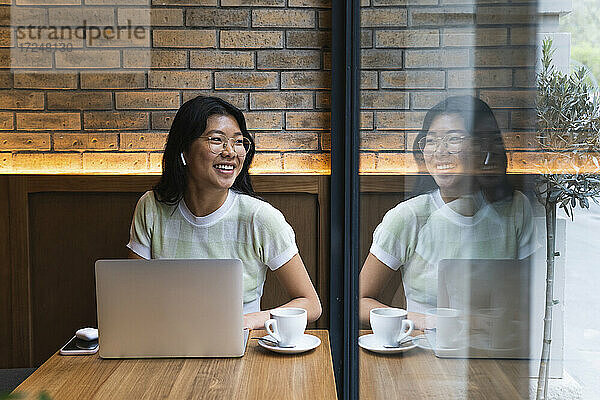 Junge Frau mit Laptop  die in der Nähe eines Glasfensters in einem Café sitzt und wegschaut