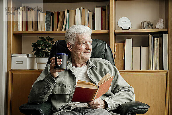 Älterer Mann mit Buch  der wegschaut und eine Tasse zu Hause hält