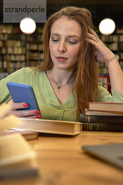 Junge Frau mit Hand im Haar  die in einer Bibliothek ein Mobiltelefon benutzt