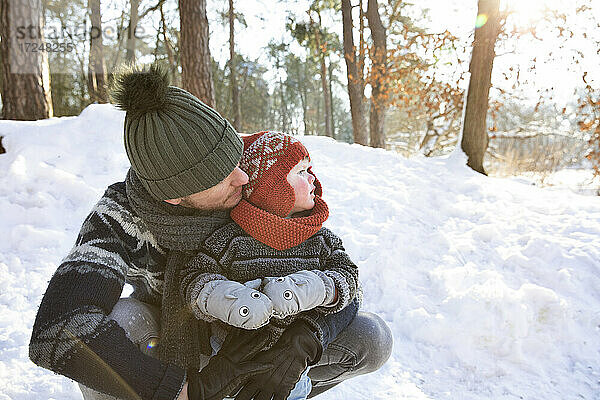 Vater mit Strickmütze  der seinen Sohn umarmt  während er im Winter im Schnee hockt