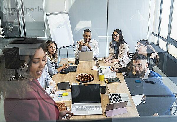 Weibliche Fachkraft erklärt Kollegen den Geschäftsplan während einer Besprechung  gesehen durch Glas in einem Coworking-Büro