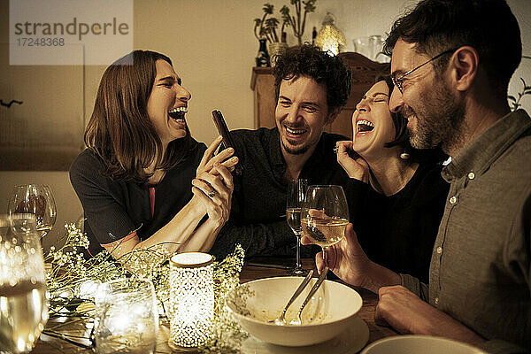 Männliche und weibliche Freunde lachen  während die Frau am Esstisch ihr Handy zeigt
