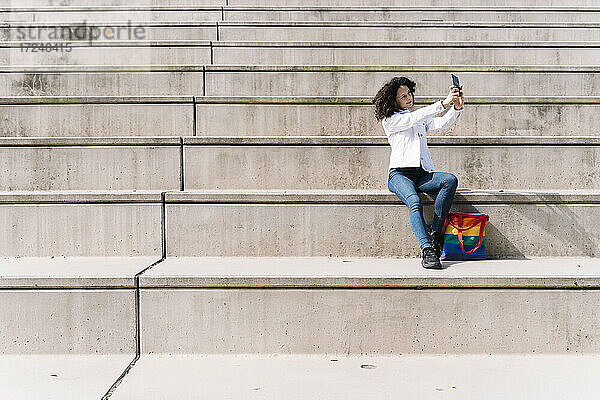 Hispanische Frau  die ein Selfie mit ihrem Smartphone macht  während sie an einem sonnigen Tag auf einer Treppe sitzt