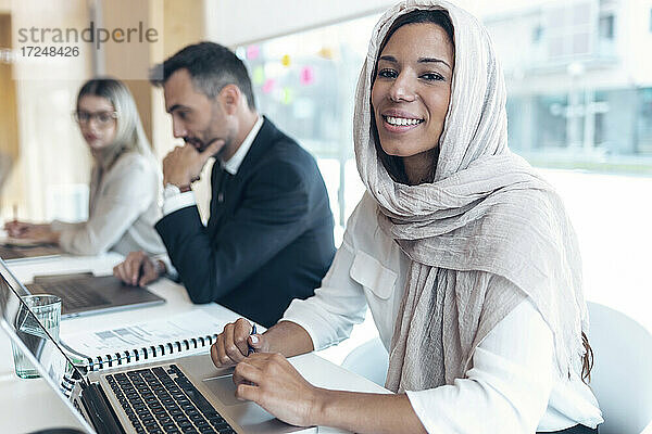 Lächelnde Geschäftsfrau mit Kopftuch am Laptop im Büro
