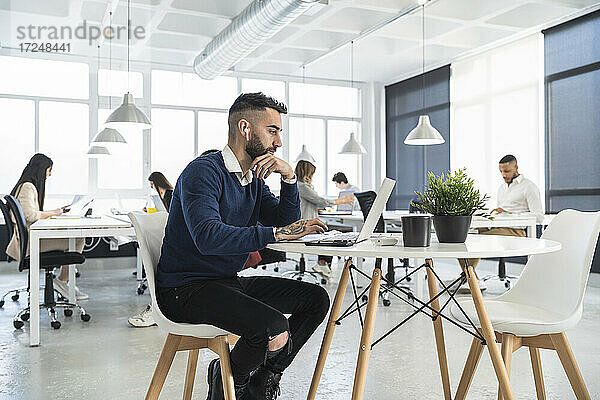 Männliche Fachkraft arbeitet an einem Laptop mit Kollegen im Hintergrund in einem Coworking-Büro