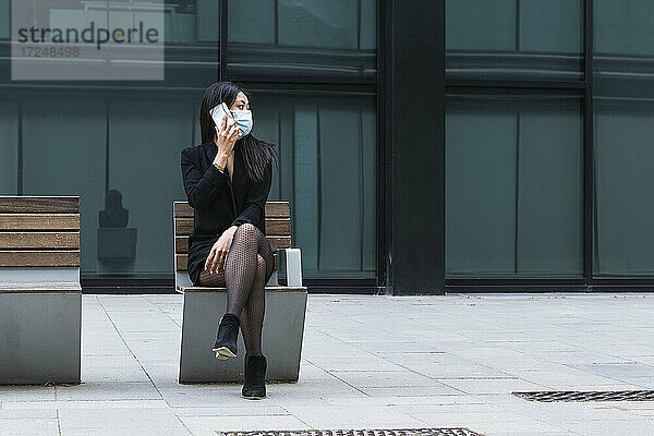 Frau mit Gesichtsmaske  die auf einer Bank sitzend mit einem Mobiltelefon spricht