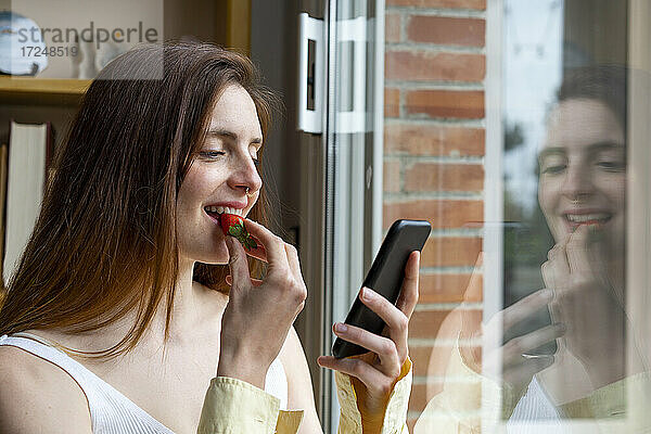 Lächelnde Frau  die ein Mobiltelefon benutzt  während sie zu Hause eine Erdbeere isst